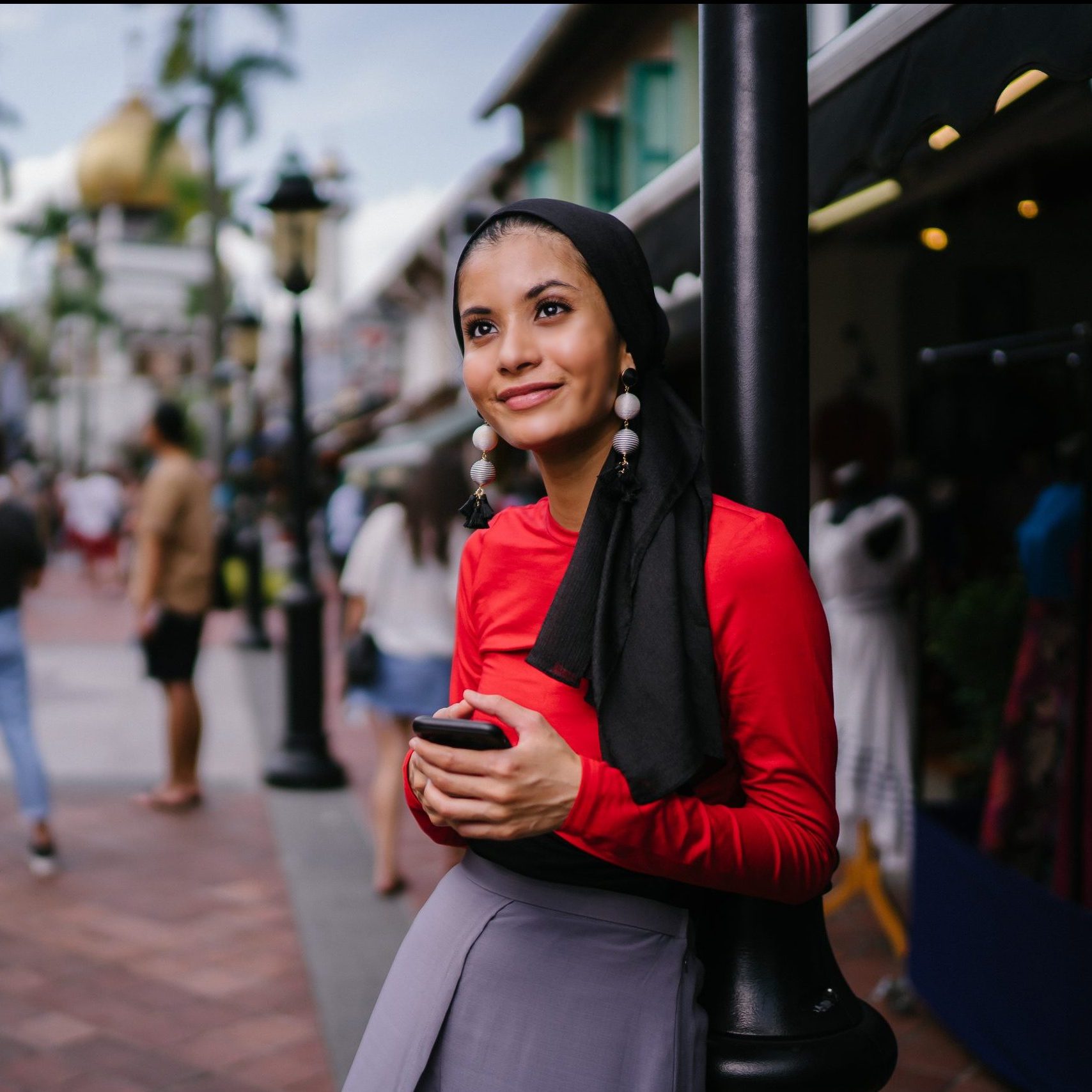A beautiful and young Muslim Middle Eastern woman texts on her smartphone as she leans against a pole on a crowded street with a mosque in the distance. She is smiling confidently.