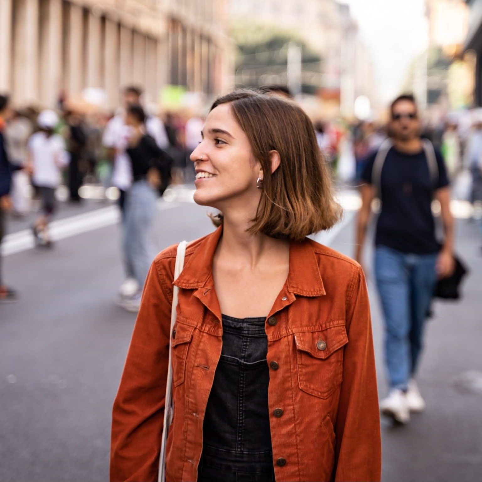 Blonde girl in the middle of the street with happy expression walking around with an orange jacket and a bag and looking to the side. Travel concept