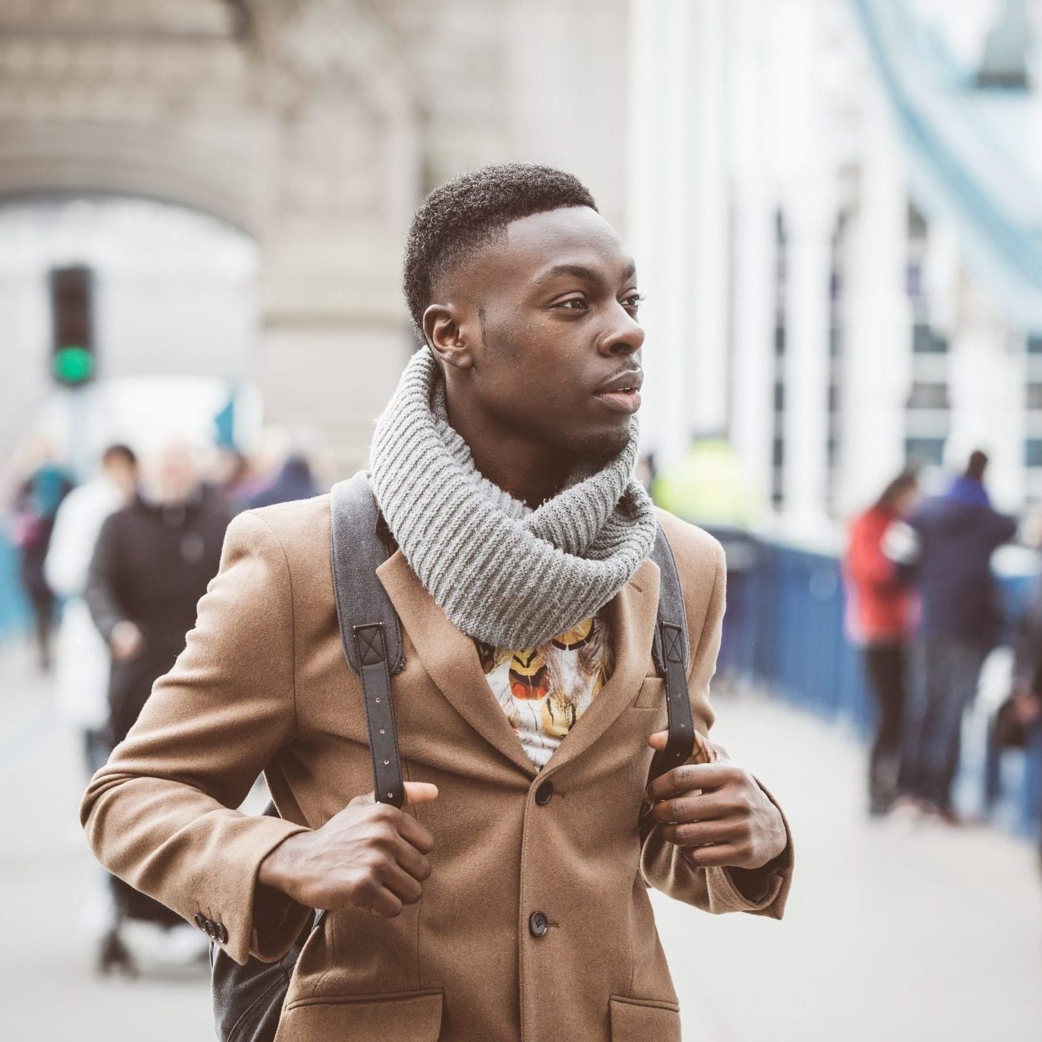 Young black man in London walking  on Tower Bridge. He wears a brown coat and light blue jeans. Traffic and other persons on background.